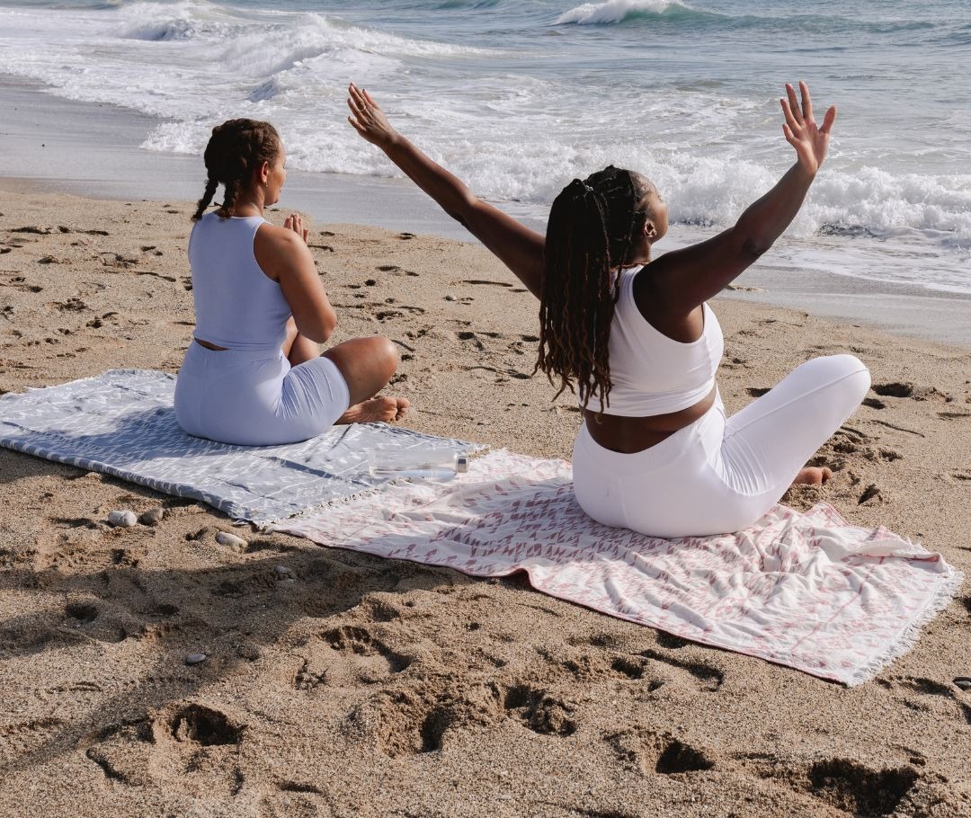 Two women practicing mindfulness and stretching on the beach near ocean waves.