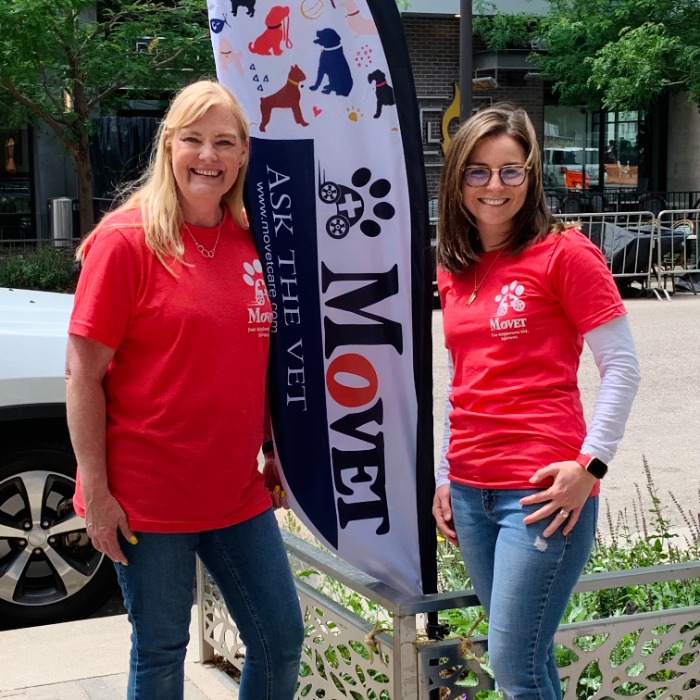 Two MoVET vets in red shirts smile beside a branded flag at an outdoor community event.